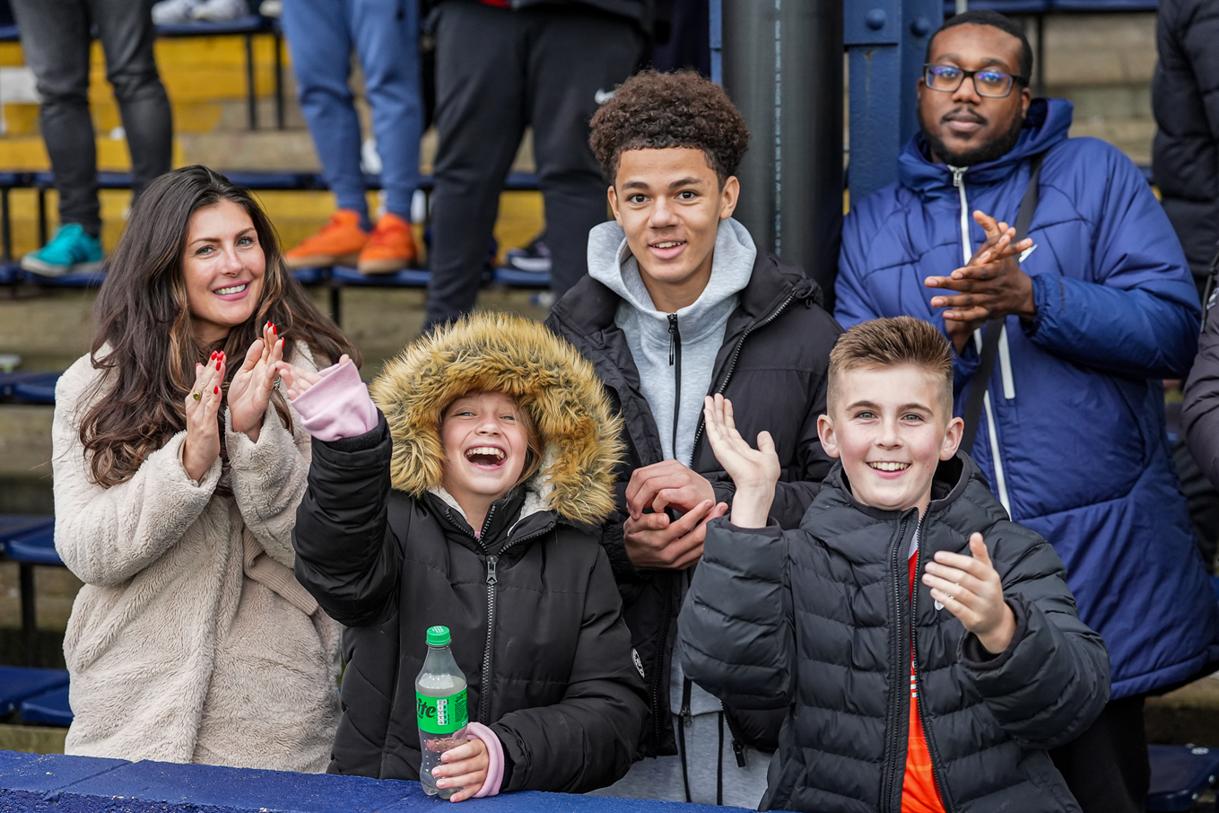 A group of Luton fans smiling and waving at the camera in the Main Stand enclosure at Kenilworth Road.